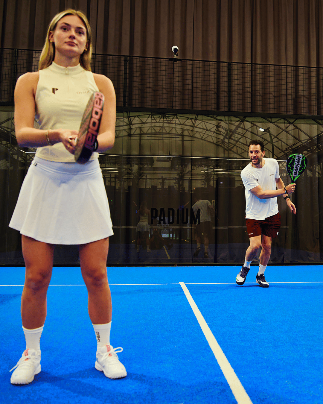 A woman in a white padel dress stands ready with her racket on a blue padel court, with a male player behind her starting the game with a serve.