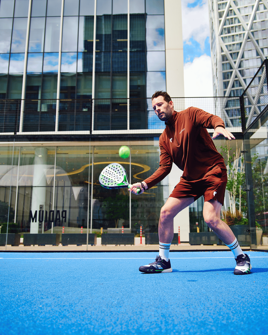A man in a rich brown outfit hits a forehand on a blue outdoor padel court, with Canary Wharf office buildings behind him.