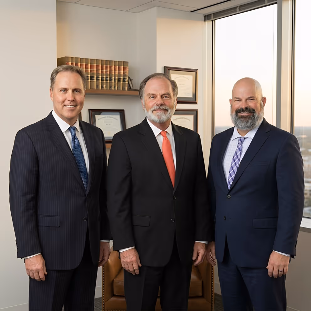 Three men in business suits standing in an office with certificates, books, and a window view.