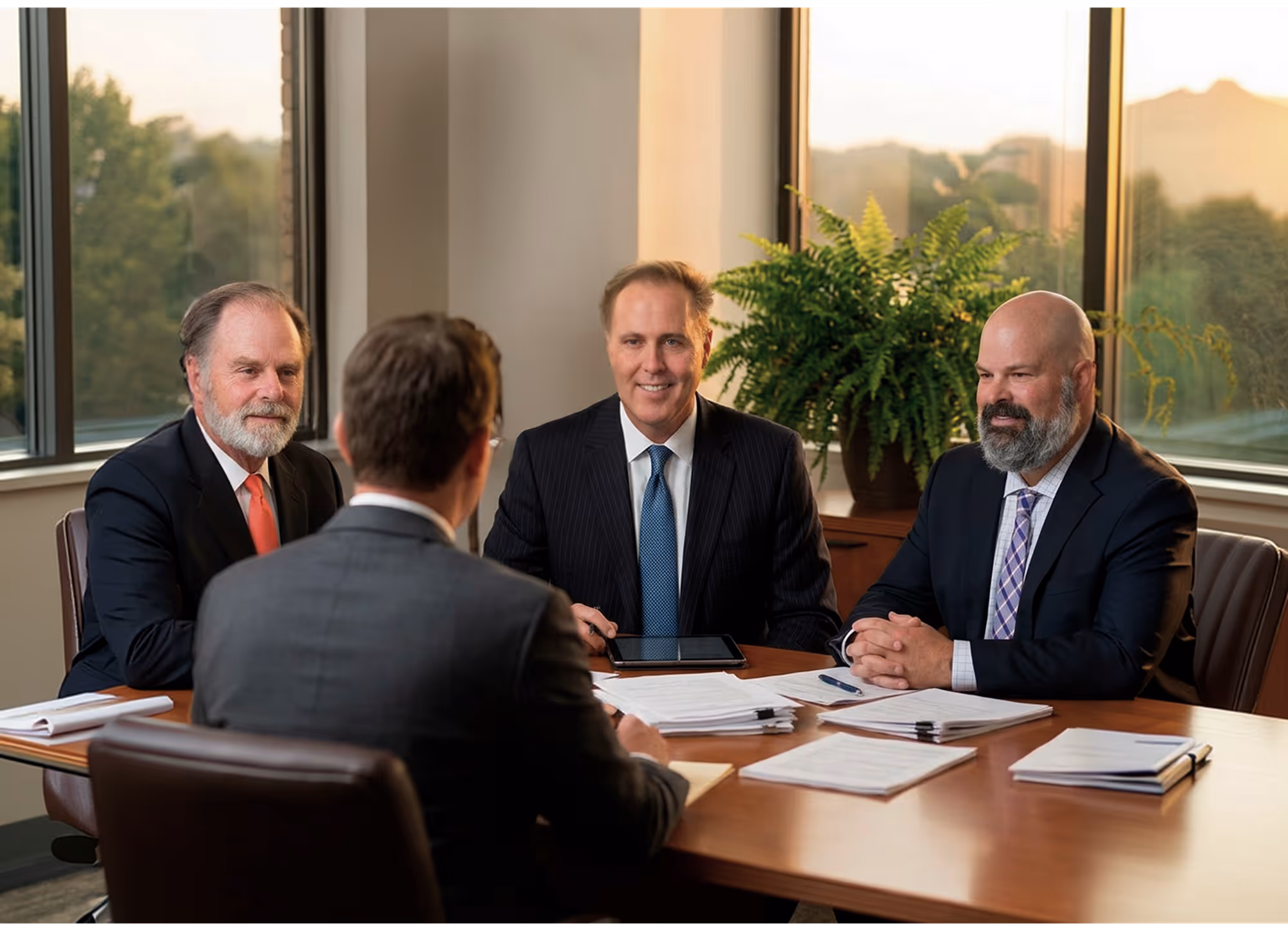 Four businessmen in suits having a meeting around a table with documents and a tablet in a bright office with large windows.