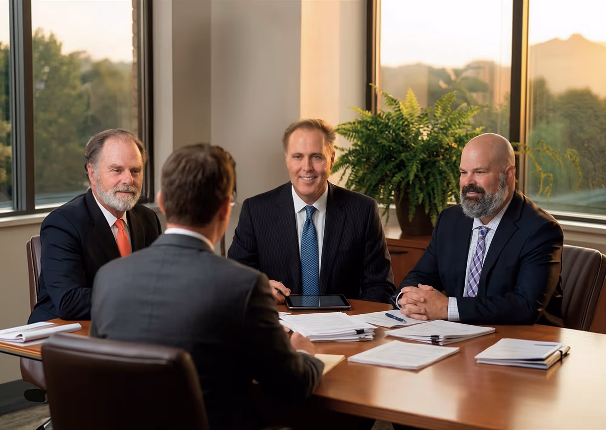 Four businessmen in suits having a meeting around a table with documents and a tablet in a bright office with large windows.