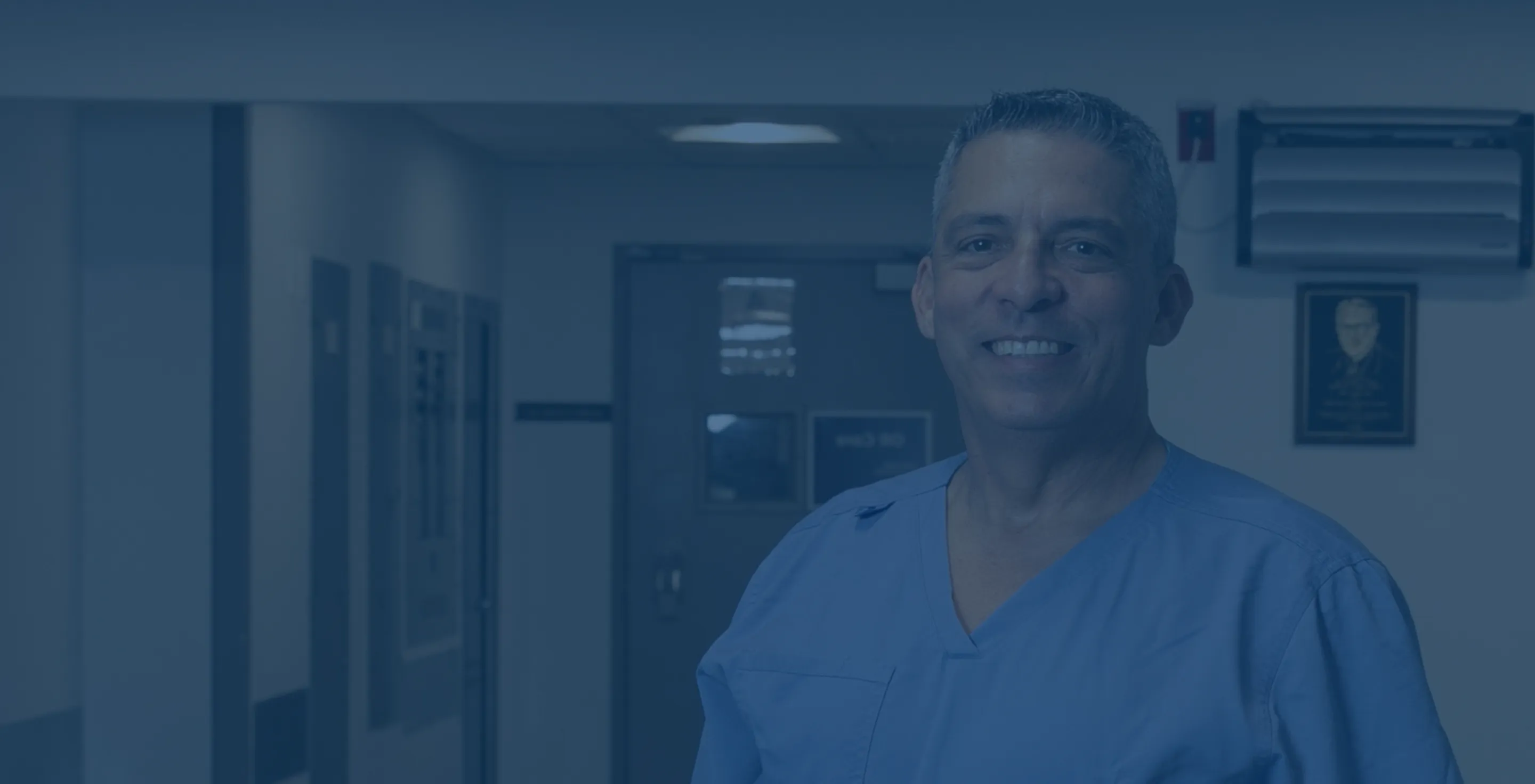 Dr. Nestor de la Cruz-Munoz wearing blue scrubs standing in a hospital corridor.