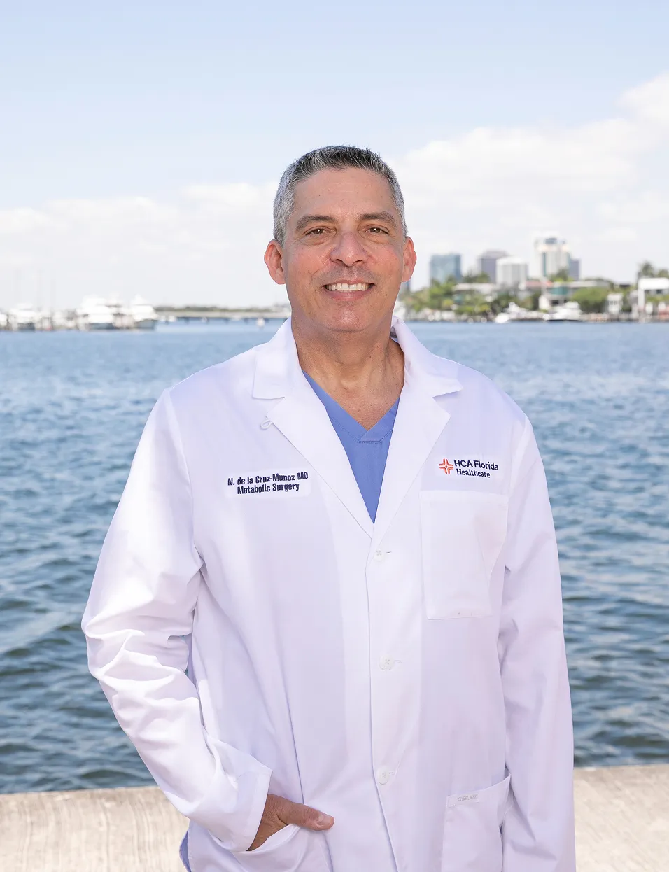 Dr. Nestor de la Cruz-Munoz wearing a white lab coat with 'N. de la Cruz-Munoz MD Metabolic Surgery' standing outdoors by a waterfront with buildings and boats in the background under a clear sky.