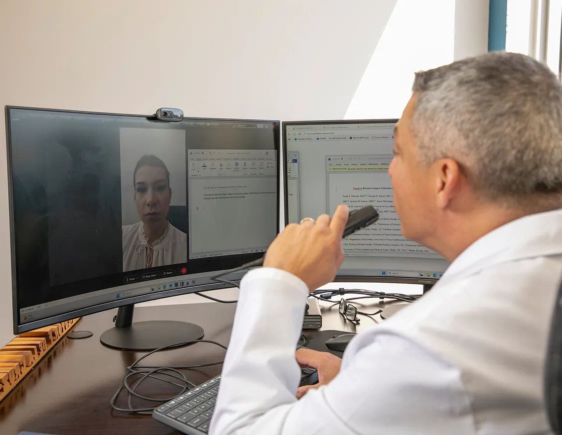 Dr. Nestor de la Cruz-Munoz in a white coat sitting at a desk participating in a video call with a woman shown on one curved monitor, while the other monitor displays a document and browser tabs.