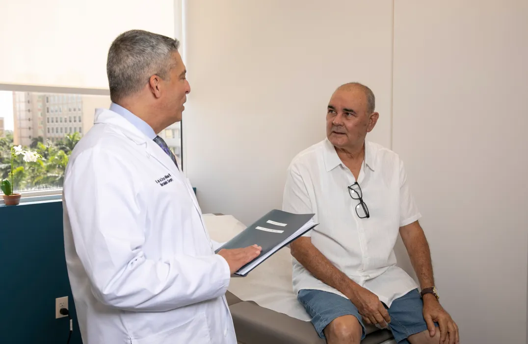 Dr. Nestor de la Cruz-Munoz in a white coat holding a folder talks to an older male patient sitting on an exam table in a medical office.