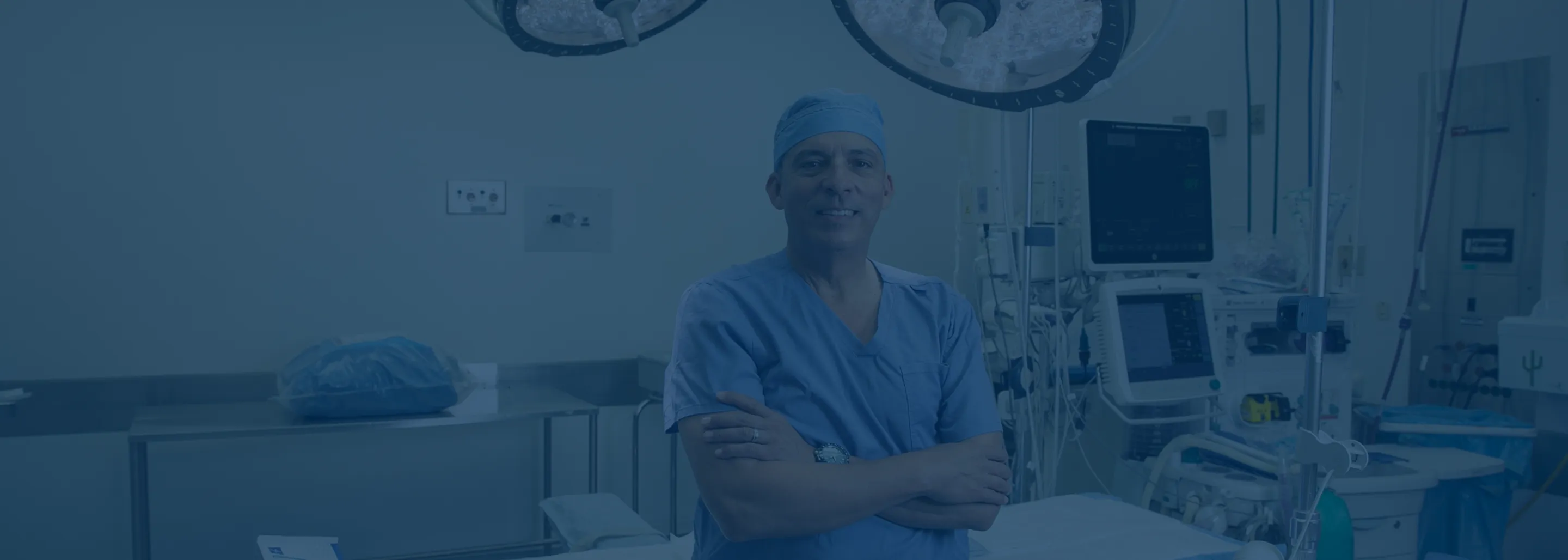A male medical professional in blue scrubs and surgical cap stands with arms crossed in a hospital operating room equipped with medical devices and monitors.