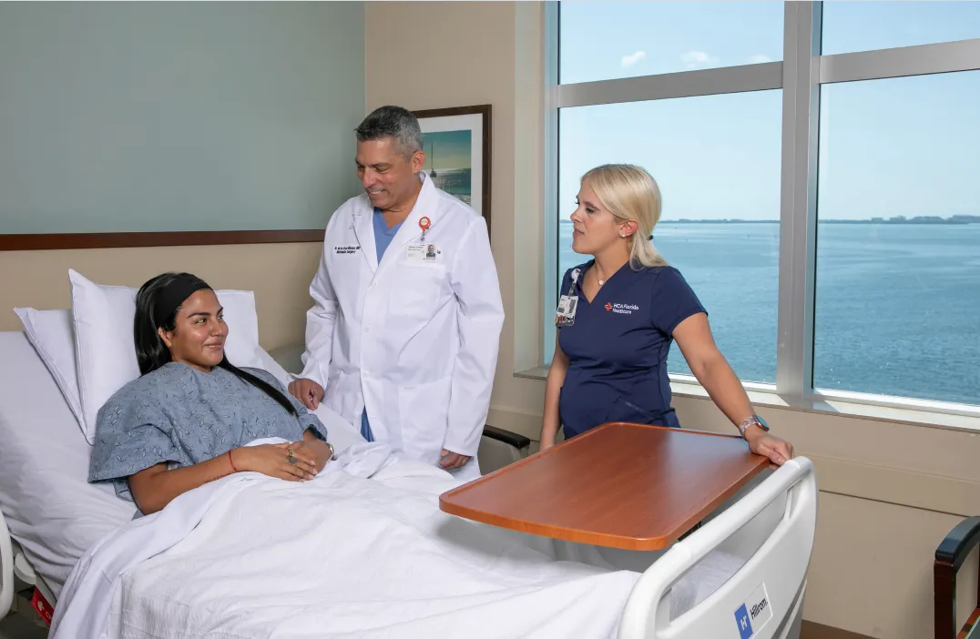 Hospital scene with a female patient lying in bed, smiling and talking with a male doctor in a white coat and a female nurse in navy scrubs, with a large window showing a body of water in the background.