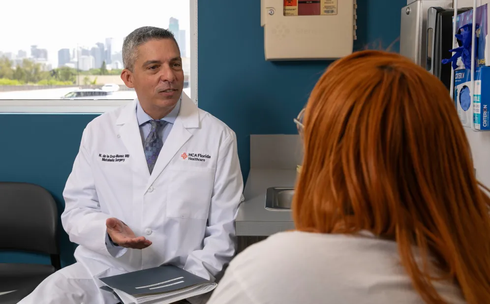 A doctor in a white coat labeled 'HCA Florida Healthcare' and 'Metabolic Surgery' is speaking to a patient with long red hair in a medical office with a window showing a cityscape.