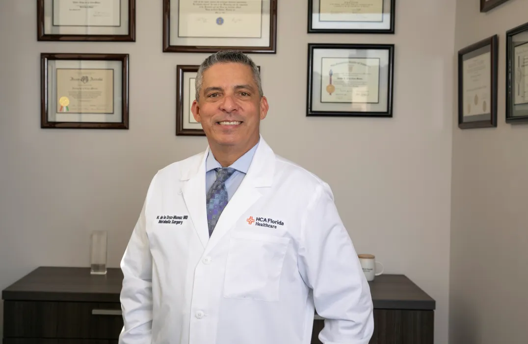 Dr. Nestor de la Cruz-Munoz wearing a white coat and a tie, standing in an office with framed certificates on the wall behind him.