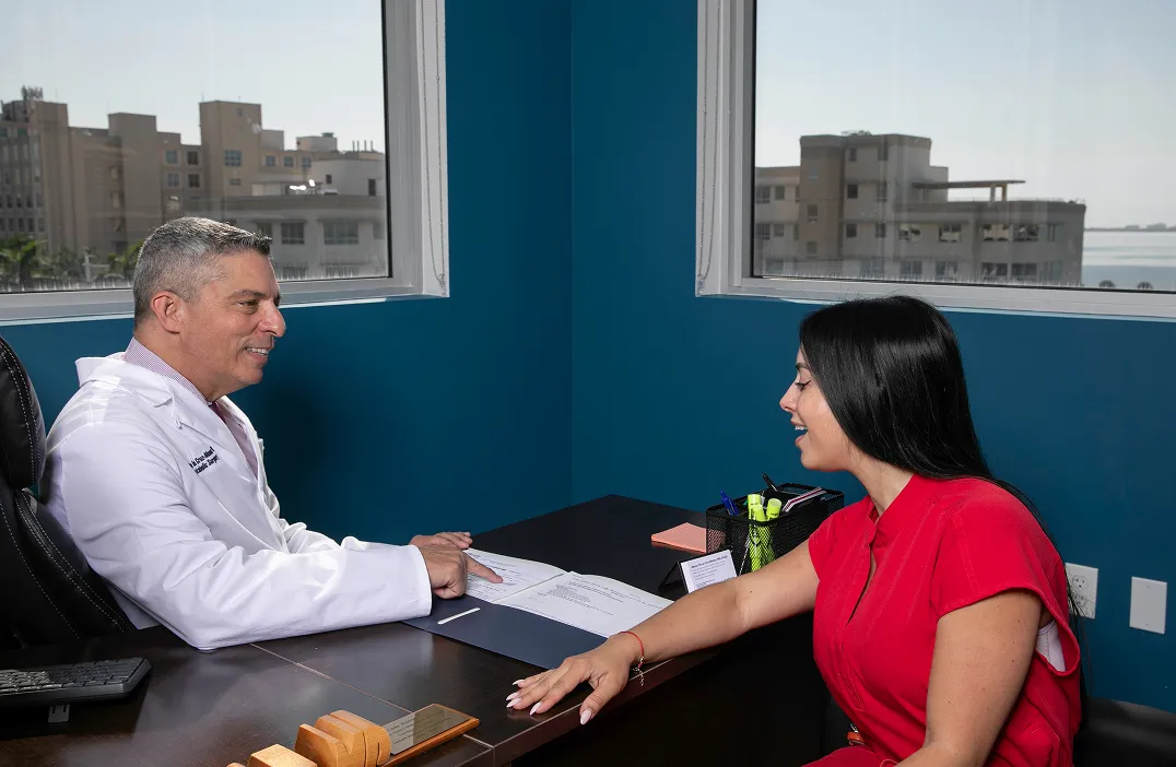 A male doctor in a white coat is sitting at a desk, smiling and pointing at a document while talking to a smiling woman in a red dress sitting across from him in an office with blue walls and large windows showing city buildings.