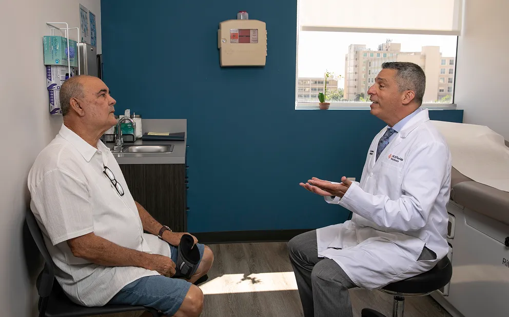 A doctor in a white coat talking to an older man seated across from him in a medical office with blue and white walls and a window showing buildings outside.