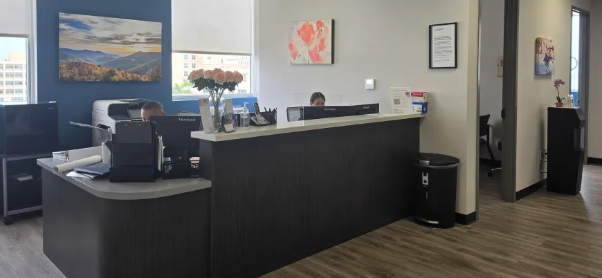 Modern office reception area with dark wood desk, two computer monitors, a vase of pink roses, wall art, and two people working behind the desk.