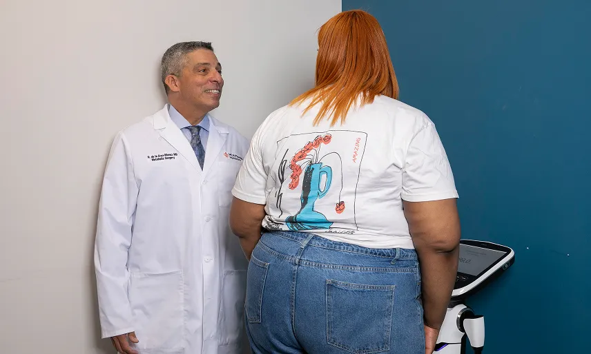 Dr. Nestor de la Cruz-Munoz in a white coat smiling and standing next to a woman with red hair wearing a white t-shirt and blue jeans, who is looking at a digital scale against a blue wall.