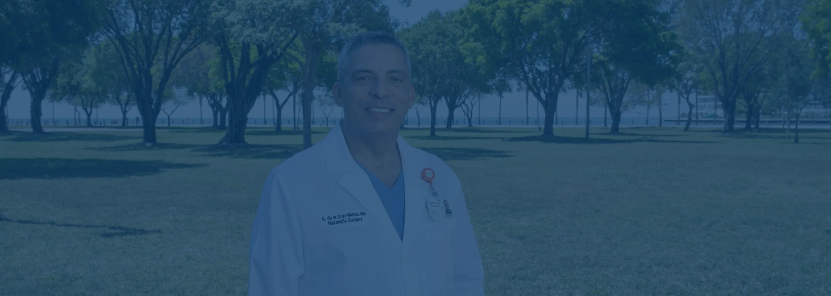 Smiling male doctor outdoors in a park wearing a white coat with a blue shirt beneath, trees and a grassy field in the background.
