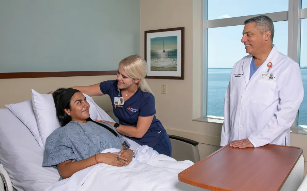 A female patient lies in a hospital bed covered with a white blanket and wearing a hospital gown, smiling at a female nurse in navy blue scrubs who is standing beside her with a comforting hand on her shoulder, while Dr. Nestor de la Cruz-Munoz in a white coat stands nearby smiling with a window showing a body of water in the background.