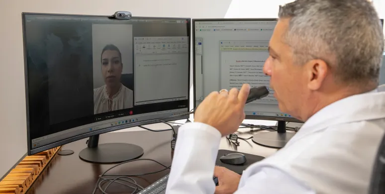 Dr. Nestor de la Cruz-Munoz sitting at a desk with two computer monitors, engaging in a video call with a woman displayed on the left screen while viewing a document on the right monitor.