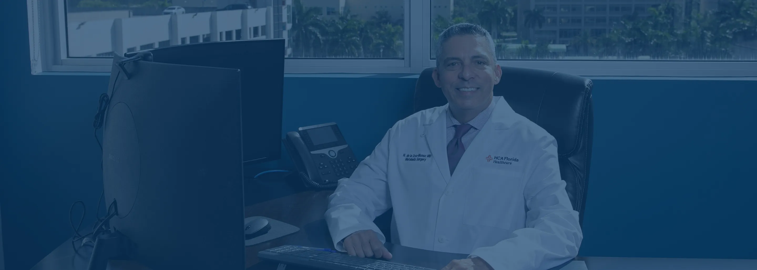 Dr. Nestor de la Cruz-Munoz in a white coat sitting at a desk with a keyboard, computer monitors, and a phone in an office with a window behind him.