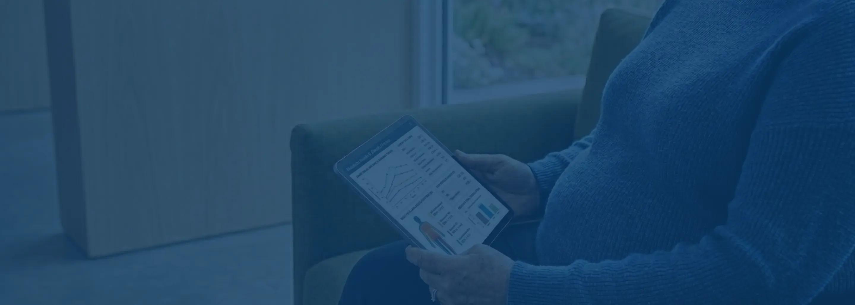 Person in a blue sweater sitting on a green armchair holding a tablet displaying health data charts and graphs.
