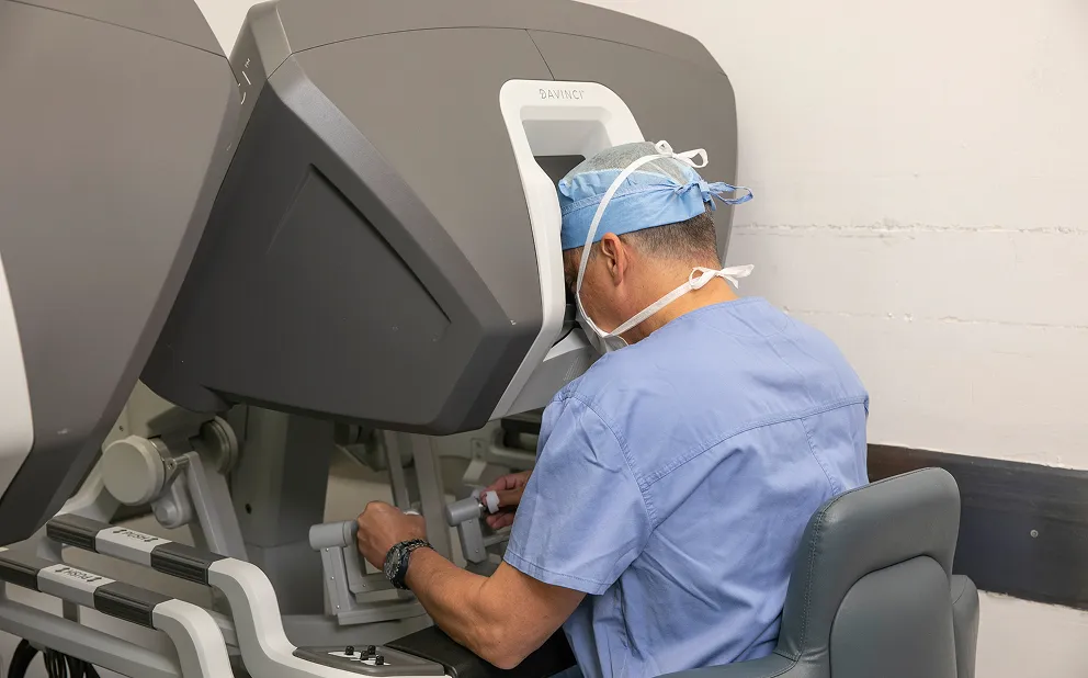 Dr. Nestor de la Cruz-Munoz in blue scrubs and cap operating a Da Vinci robotic surgery console, seated and looking into the viewfinder while manipulating controls with both hands.