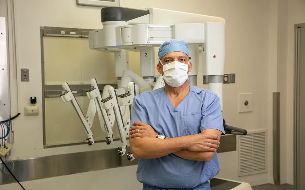 Dr. Nestor de la Cruz-Munoz in blue scrubs, surgical cap, and face mask standing with arms crossed in front of a robotic surgical system with multiple articulated arms in a hospital operating room.