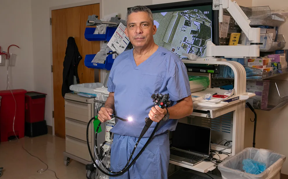 Dr. Nestor de la Cruz-Munoz in blue scrubs holding a lit endoscope in a clinical exam room with medical equipment and a monitor displaying an internal image behind him.