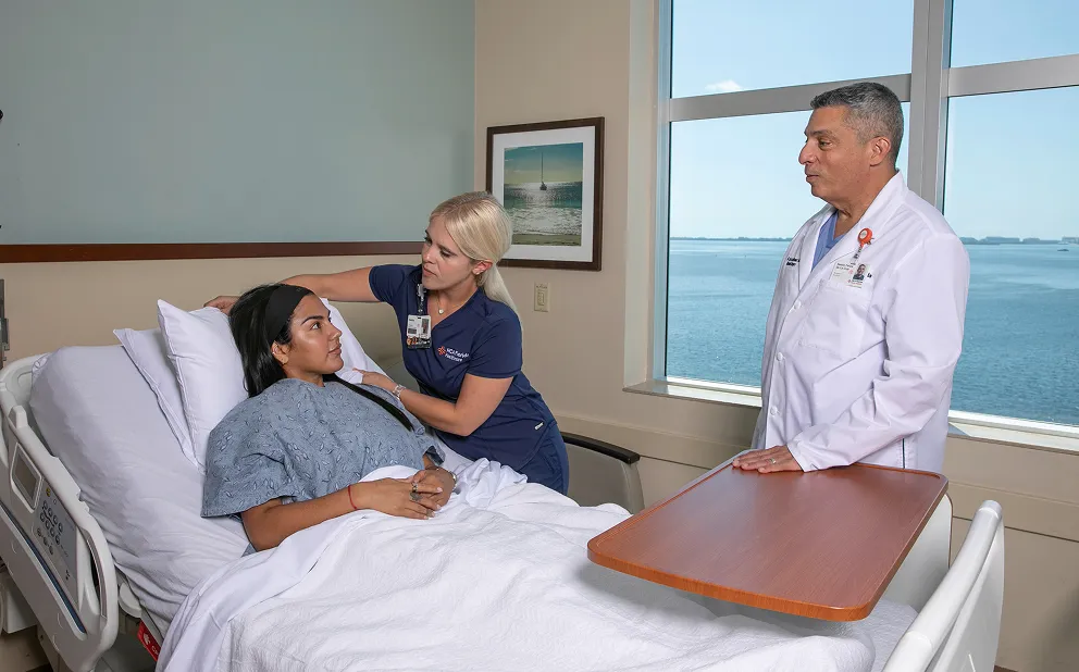 Hospital patient lying in a bed covered with white linens, attended by a female nurse in navy blue scrubs who is comforting her, whileDr. Nestor de la Cruz-Munoz in a white coat stands nearby by a window overlooking water.
