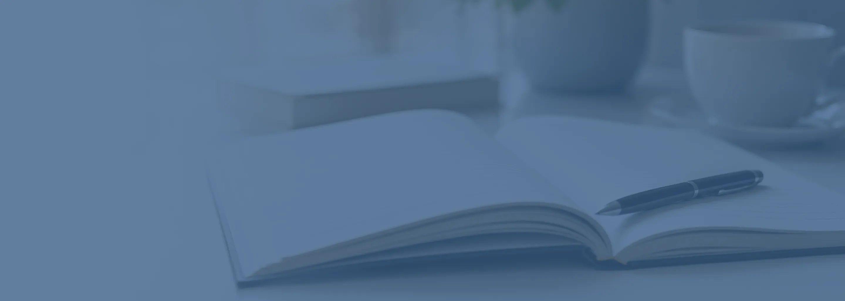 Open notebook with a pen resting on its pages, a closed book in the background, and a cup on a saucer on a desk.