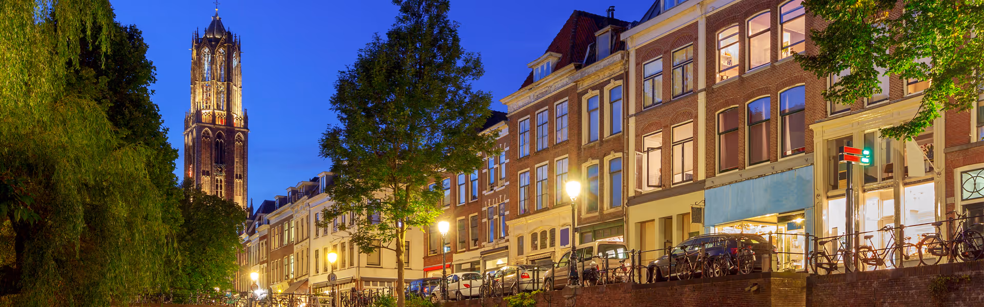 View of a lit-up church tower at dusk with trees and traditional European brick buildings along a canal street with parked cars and bicycles.