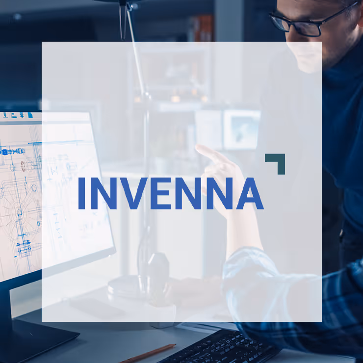 Two people working together in an office, one pointing at a computer screen displaying a technical blueprint.
