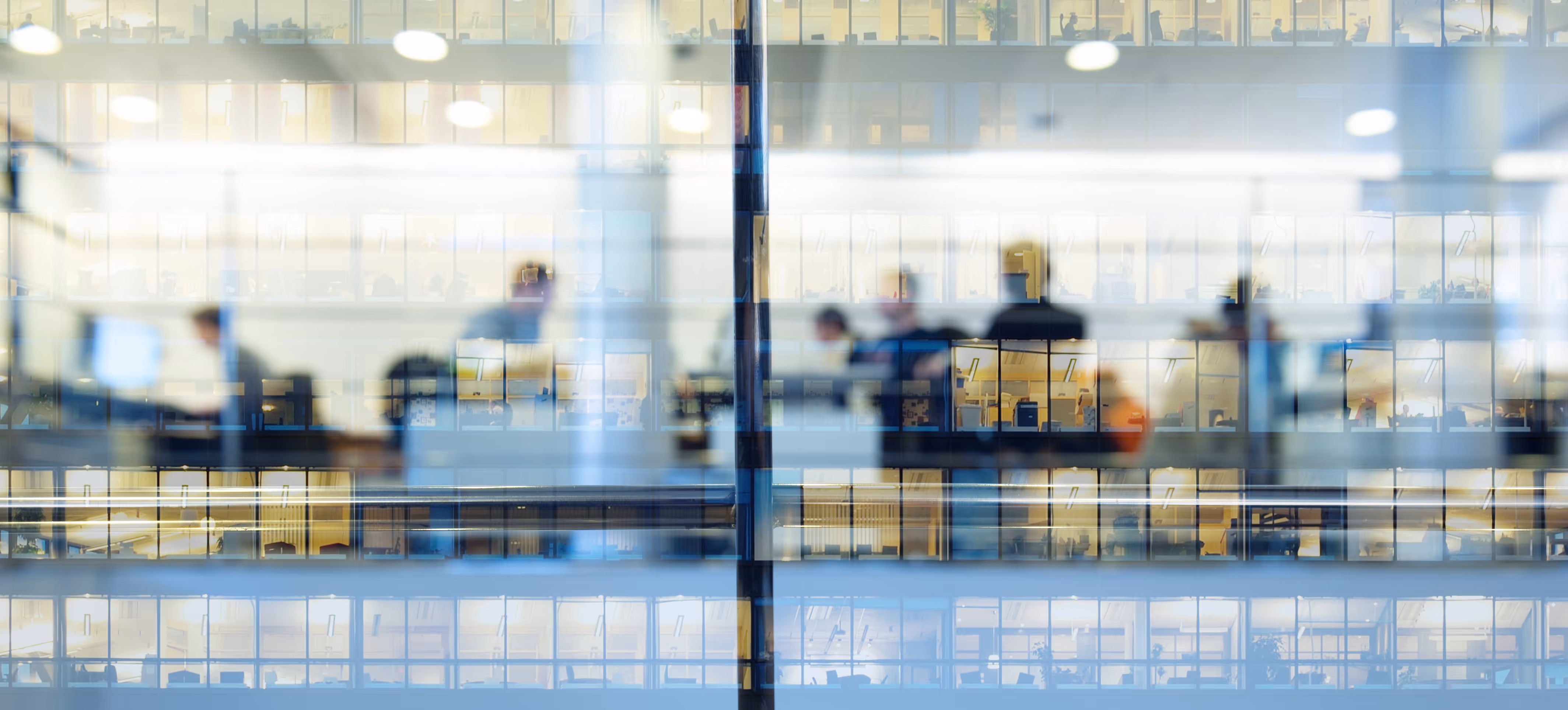 Blurred silhouettes of office workers inside a modern glass-walled office building at night.