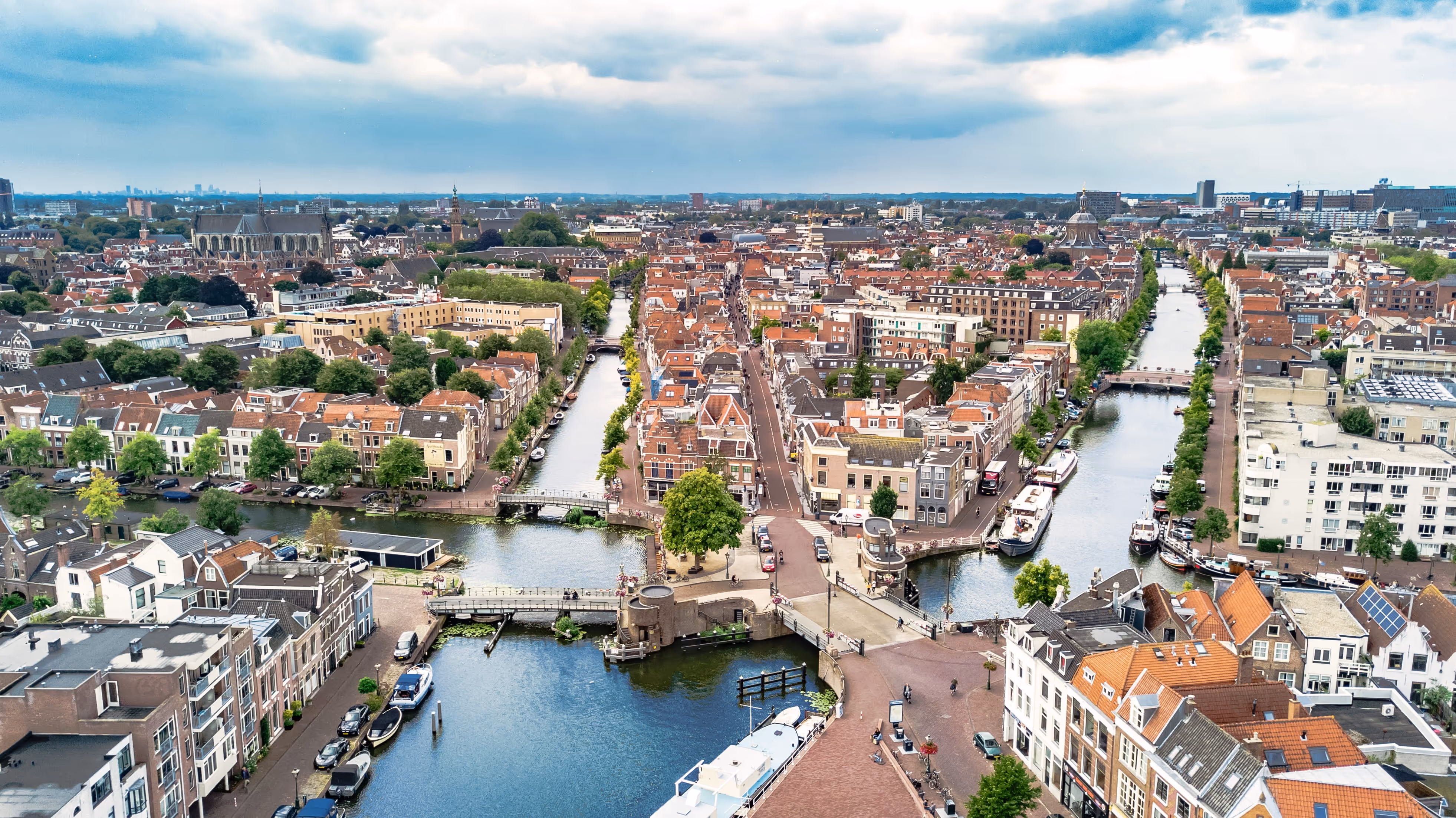 Aerial view of a Dutch city with canals, bridges, and traditional orange-roofed buildings under a partly cloudy sky.