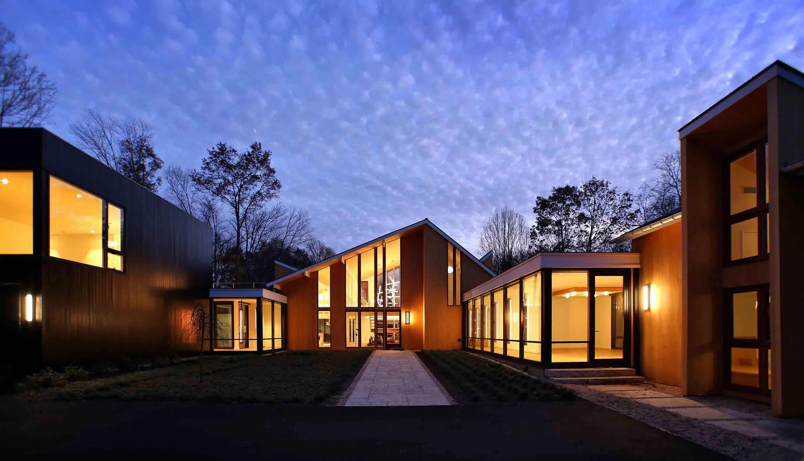 Modern house with large windows illuminated at dusk under a cloudy sky surrounded by trees.