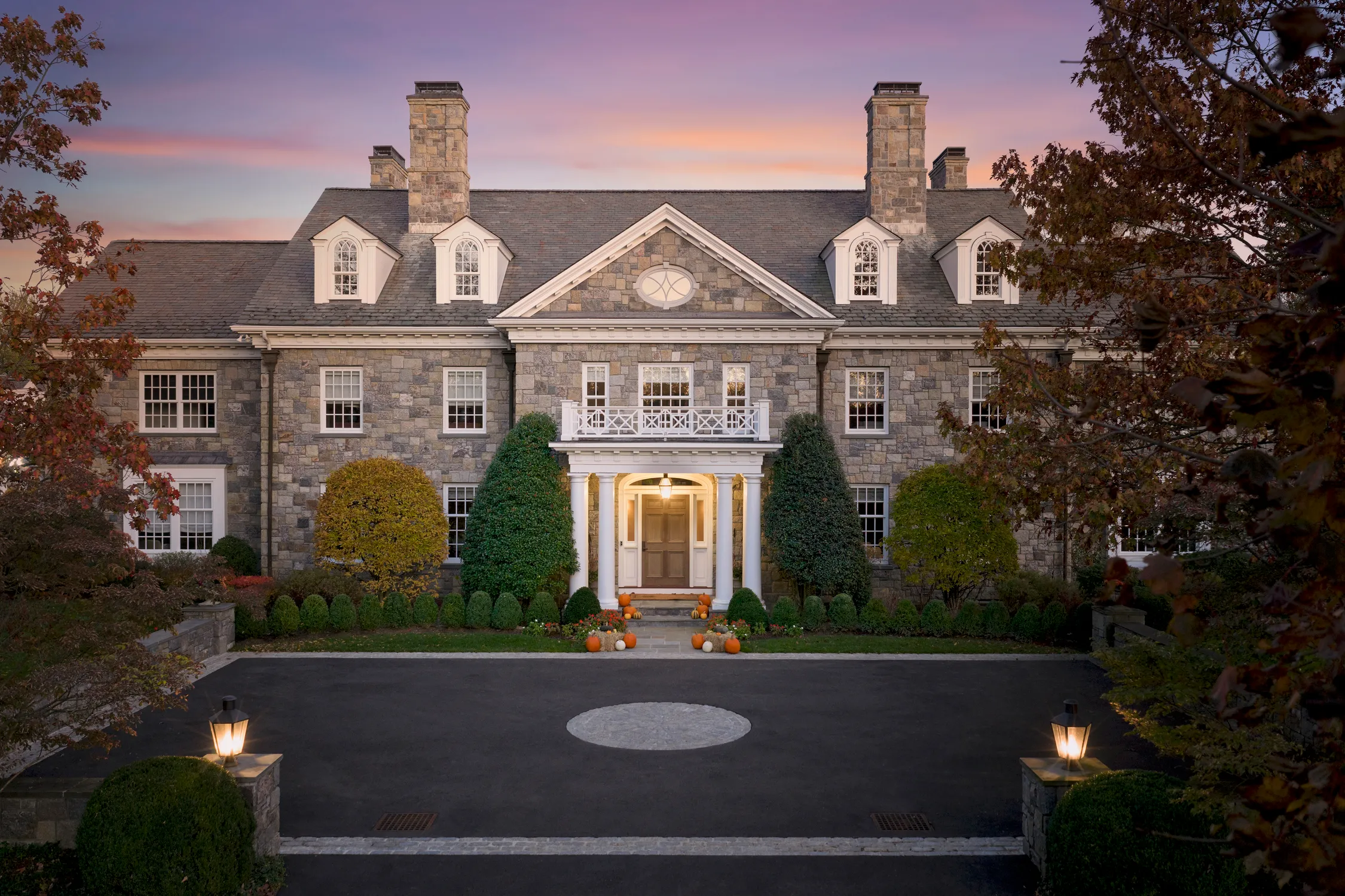 Stone mansion at dusk with symmetrical windows, white columns at the entrance, and autumn pumpkins decorating the front steps.