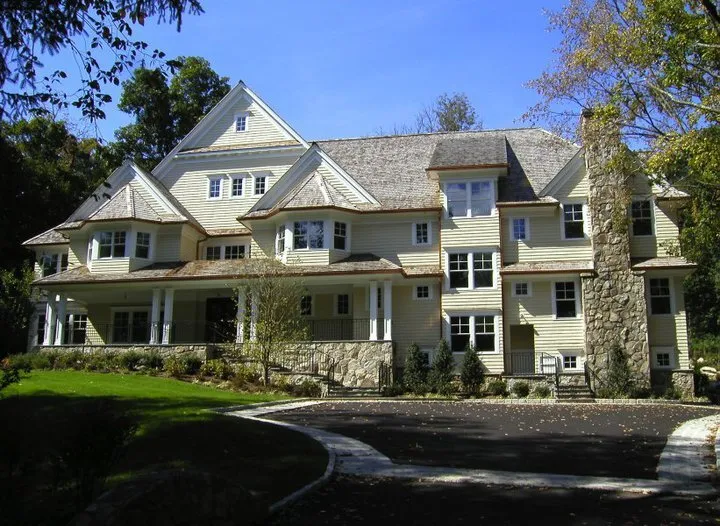 Large multi-story beige house with multiple gables, a stone chimney, wraparound porch, and surrounding trees under a blue sky.