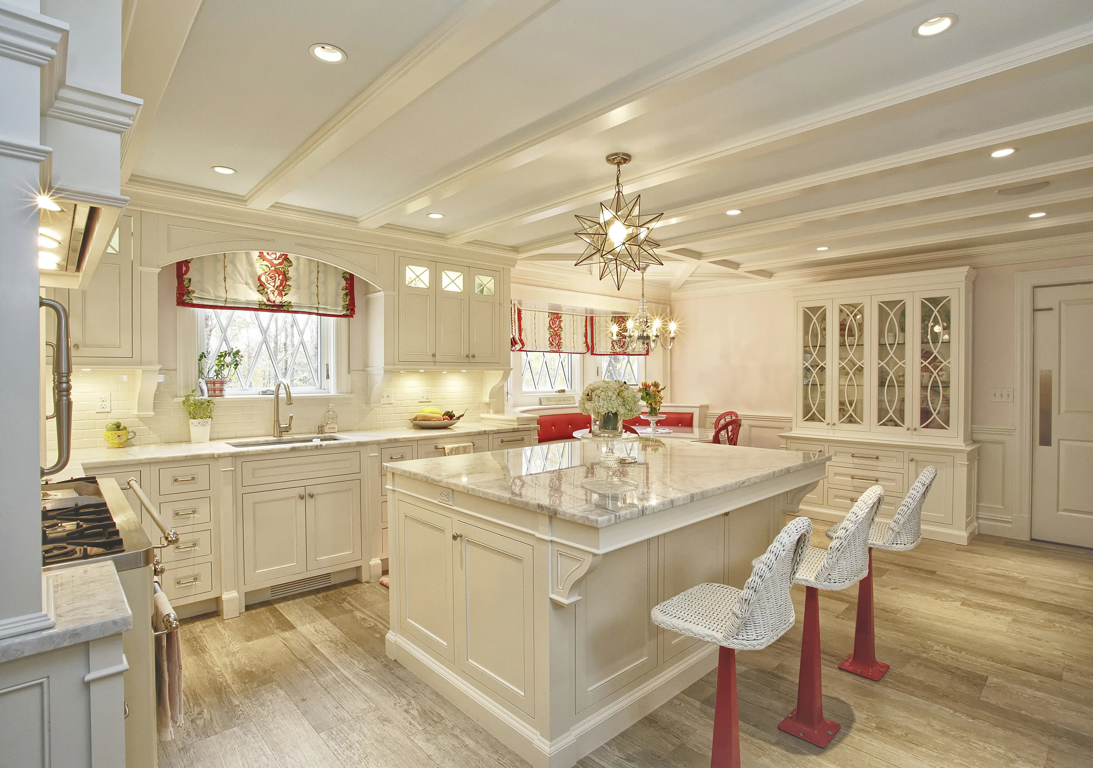 Bright kitchen with cream cabinets, marble island, three white wicker bar stools with red bases, and floral valances on windows.