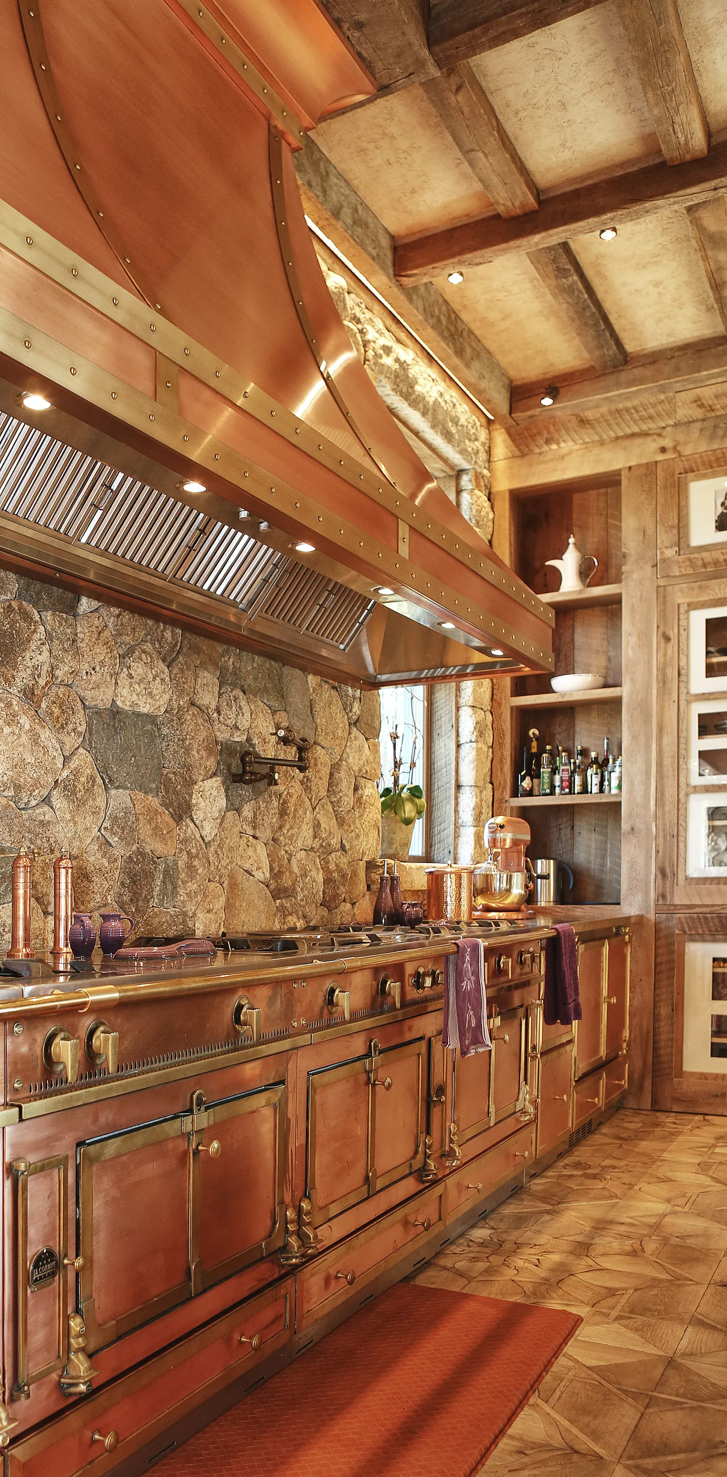Rustic kitchen with a large copper stove and hood, stone wall backsplash, wooden shelves with bottles, and wooden ceiling beams.