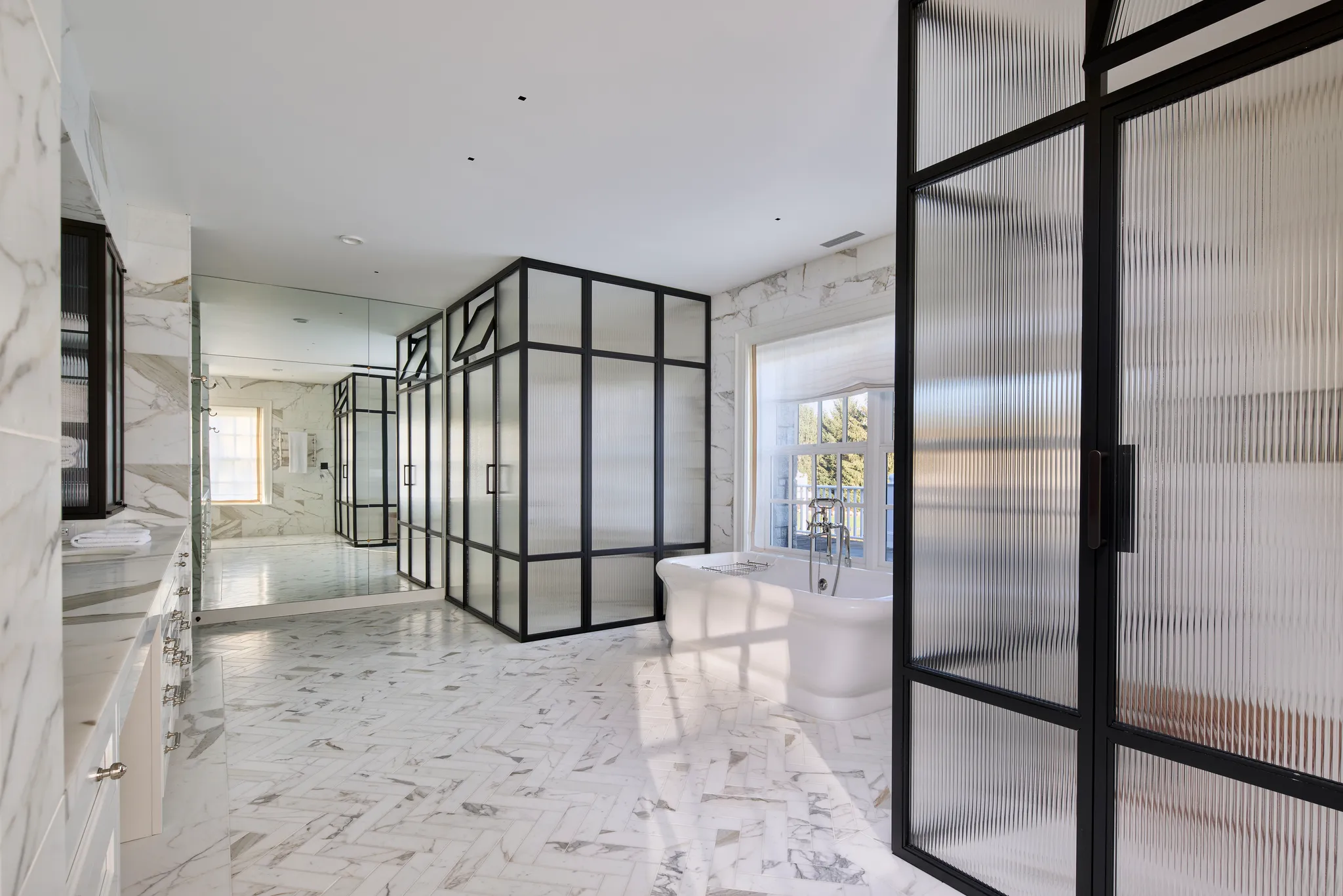 Spacious modern bathroom with white marble floors and walls, a freestanding bathtub near a window, and black-framed frosted glass shower enclosures.