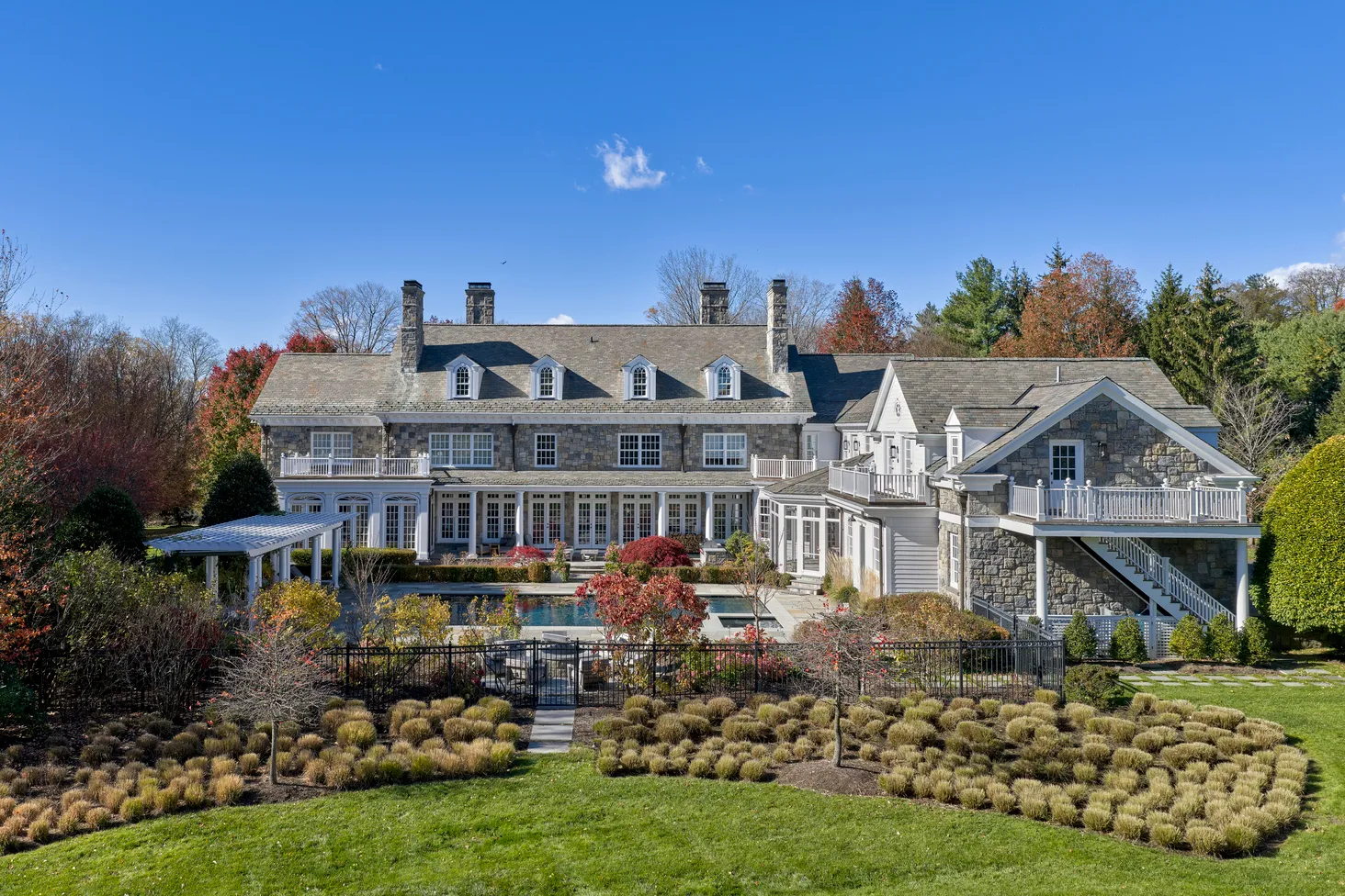Large stone and white siding house with multiple chimneys, surrounded by landscaped gardens and a pool in the backyard under a clear blue sky.