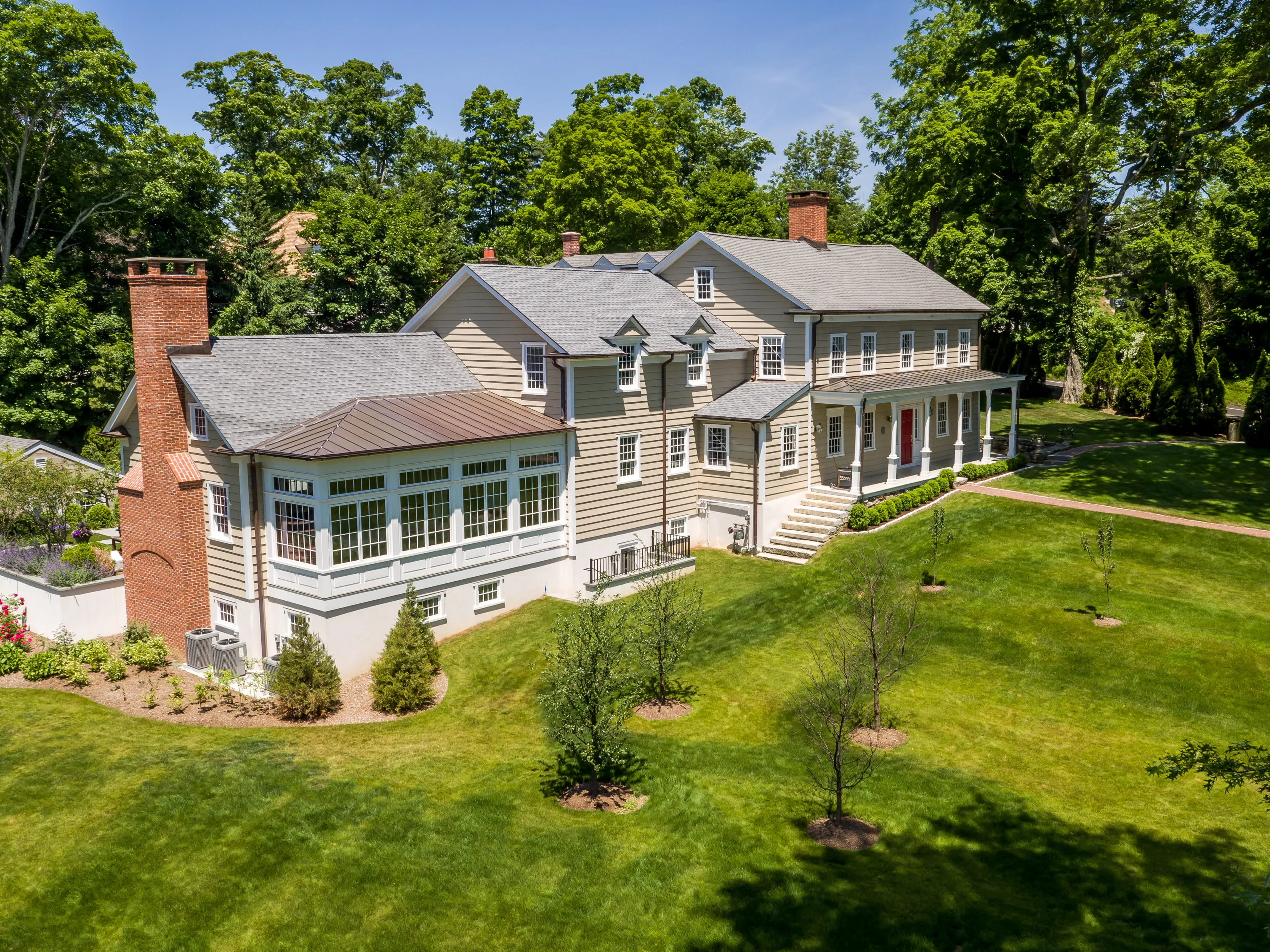 Large two-story beige house with multiple chimneys, a porch with white columns, and a well-maintained green lawn with young trees.