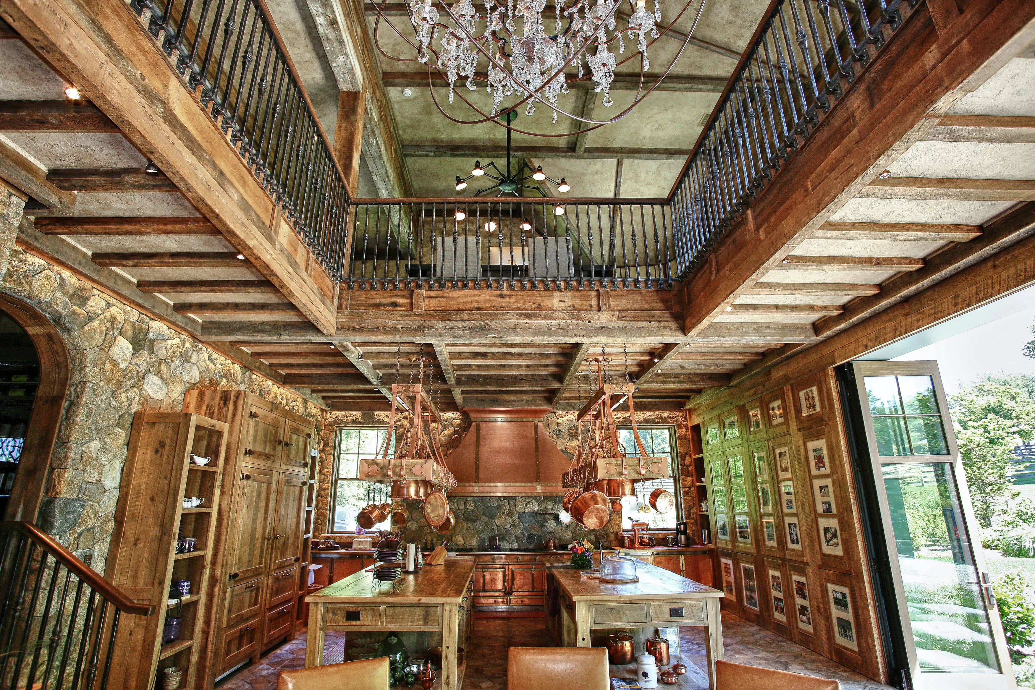 Rustic kitchen with stone walls, copper pots hanging above wooden islands, and an open door showing greenery outside.