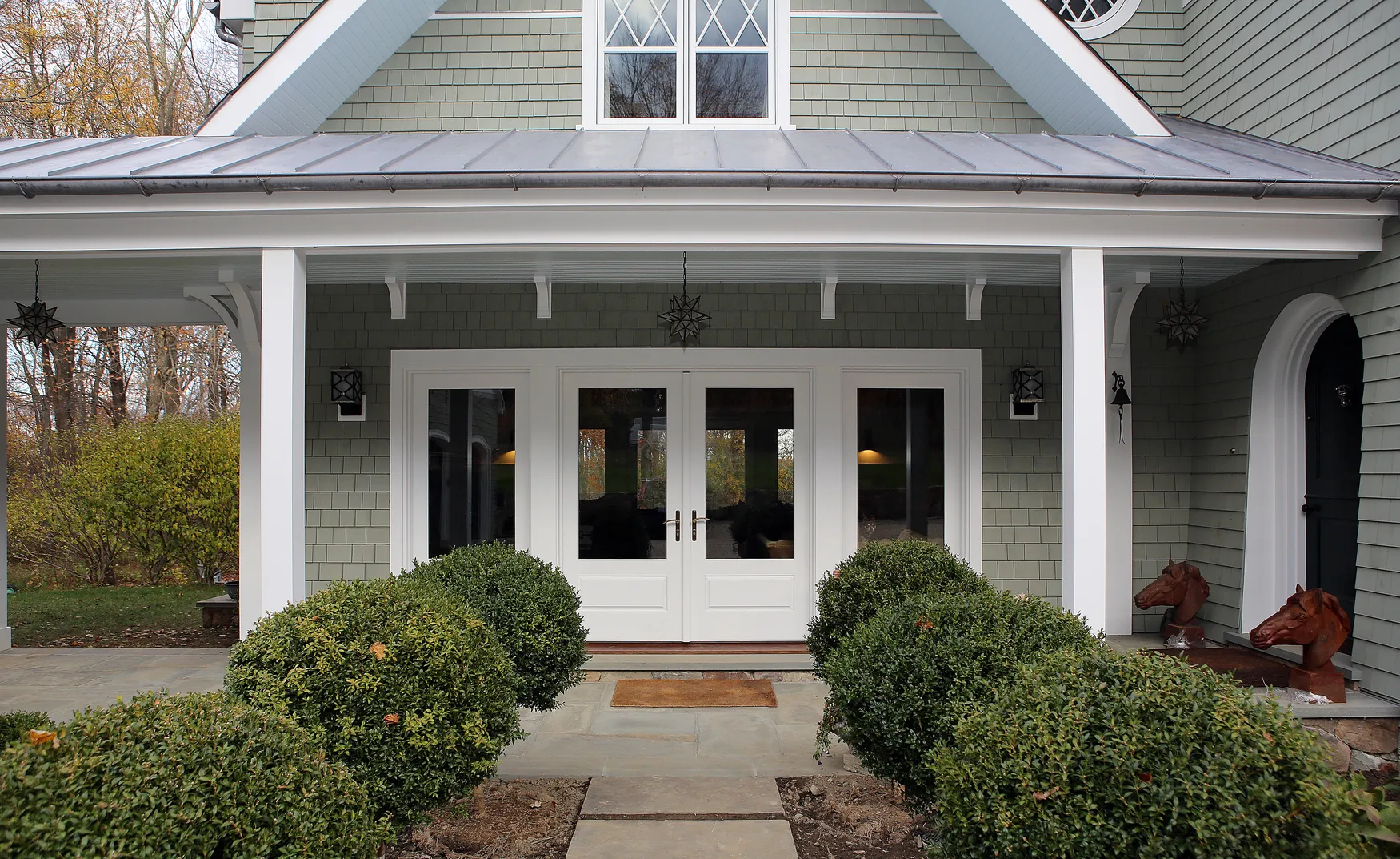 Front porch of a house with trimmed round bushes along the walkway and white double doors under a covered roof.