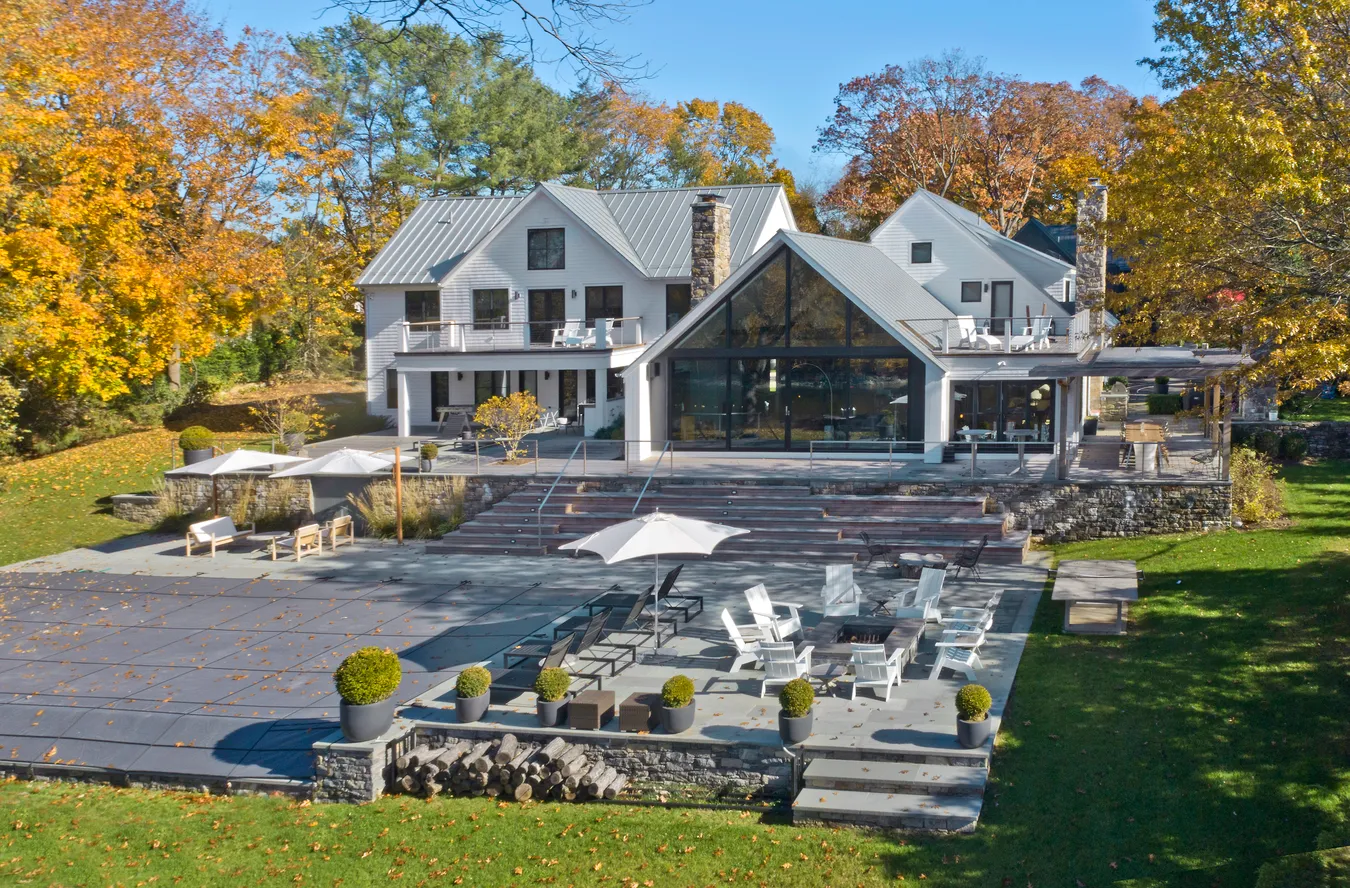 Large modern house with white exterior, metal roof, outdoor patio seating, umbrellas, fire pit, and surrounded by autumn trees.