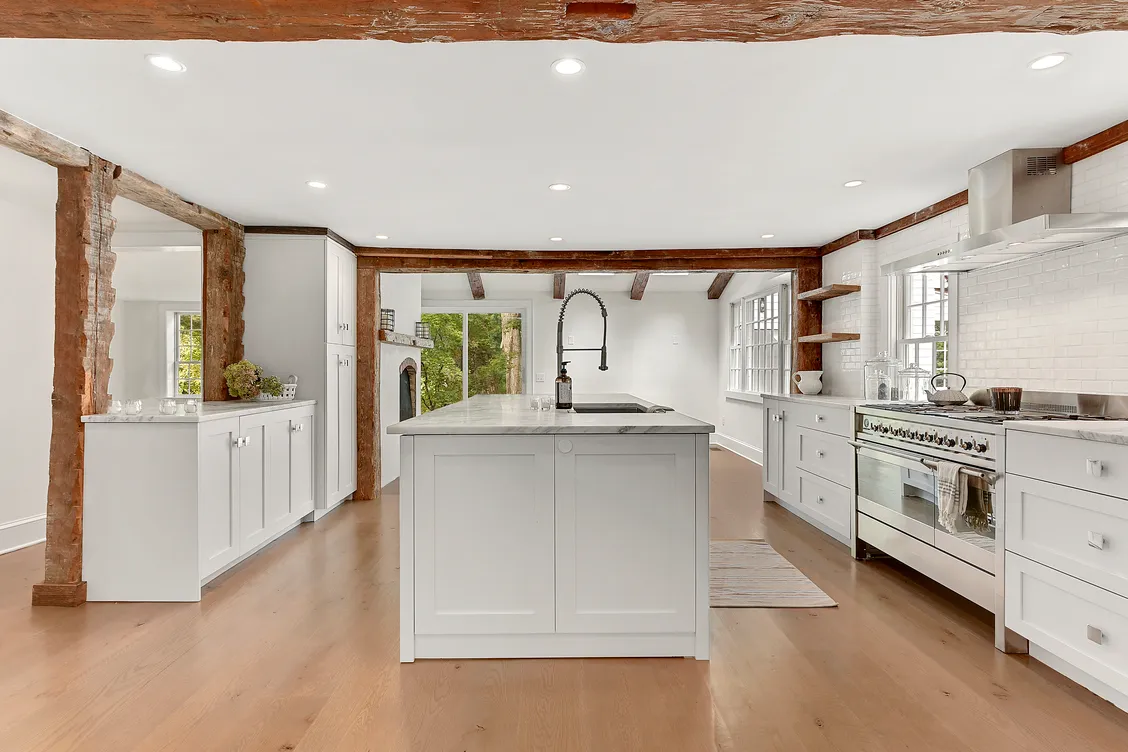 Spacious modern kitchen with white cabinetry, central island with sink and faucet, wooden beams, and stainless steel stove and hood.