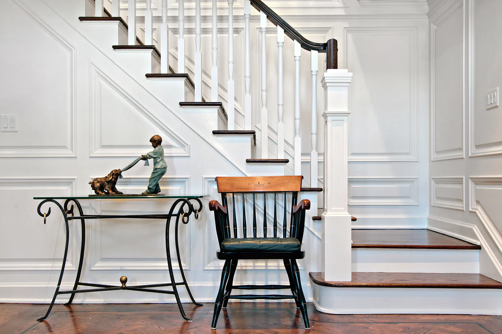 Black wooden chair with a brown backrest and black leather seat placed by white staircase with dark wood steps and white panel walls, next to a glass table with a bronze sculpture of a boy playing tug-of-war with a dog.