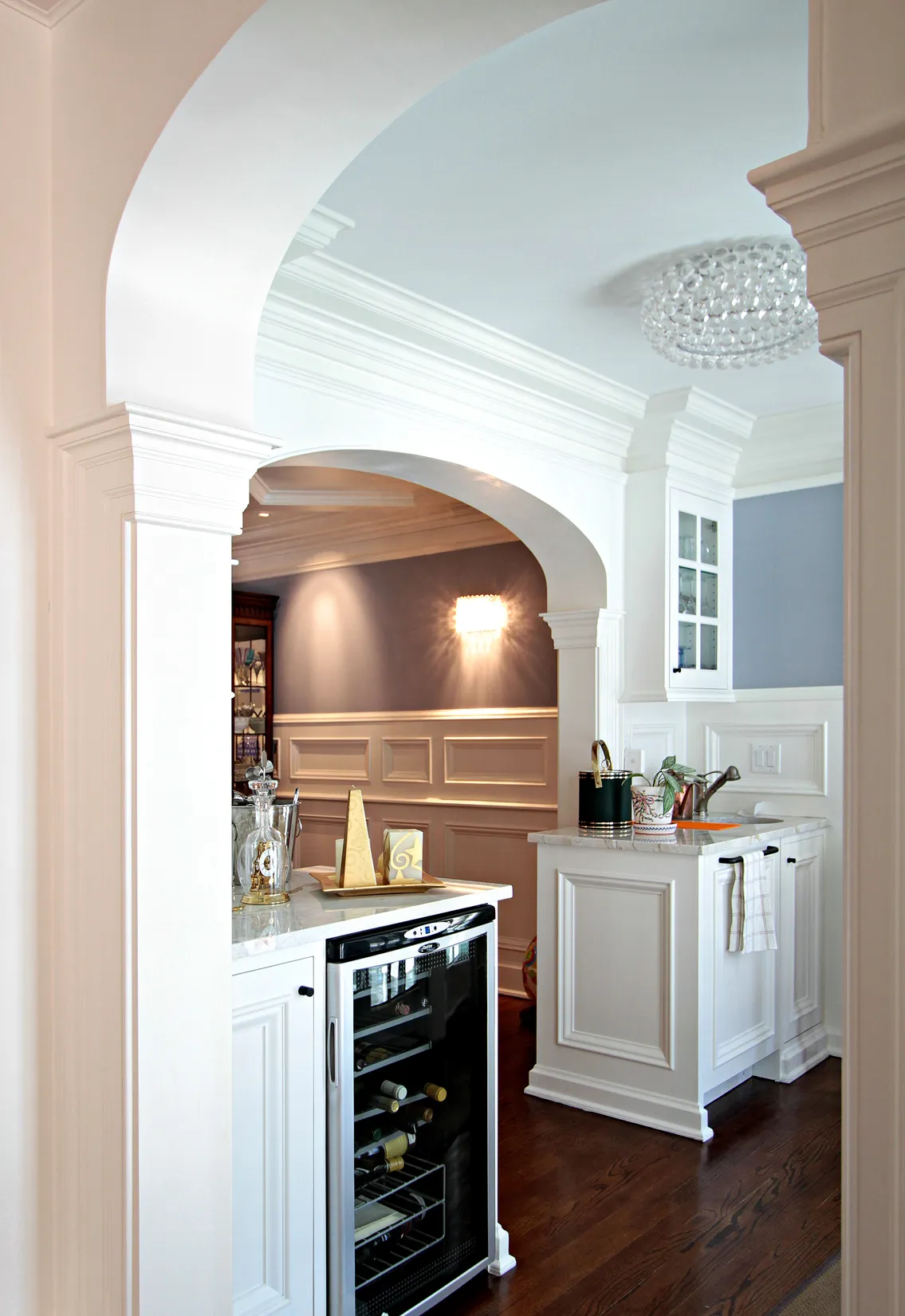 Elegant white kitchen with white cabinetry, marble countertops, built-in wine cooler, and an arched entryway.