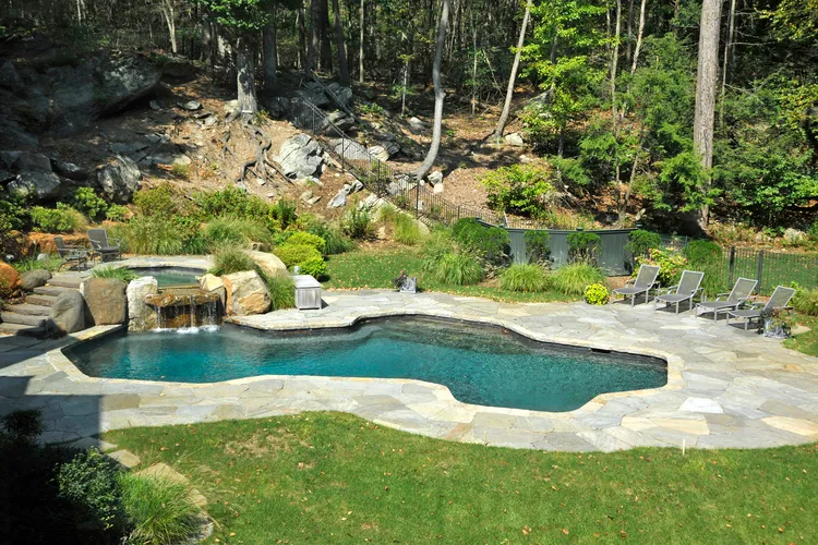 Swimming pool with attached hot tub and rock waterfall surrounded by stone patio and forested landscape.