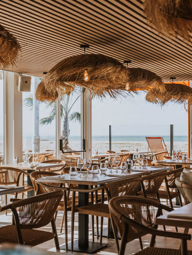 Salle de restaurant avec tables dressées, chaises en osier, lampes en paille suspendues et vue sur la plage et la mer à travers une grande baie vitrée.