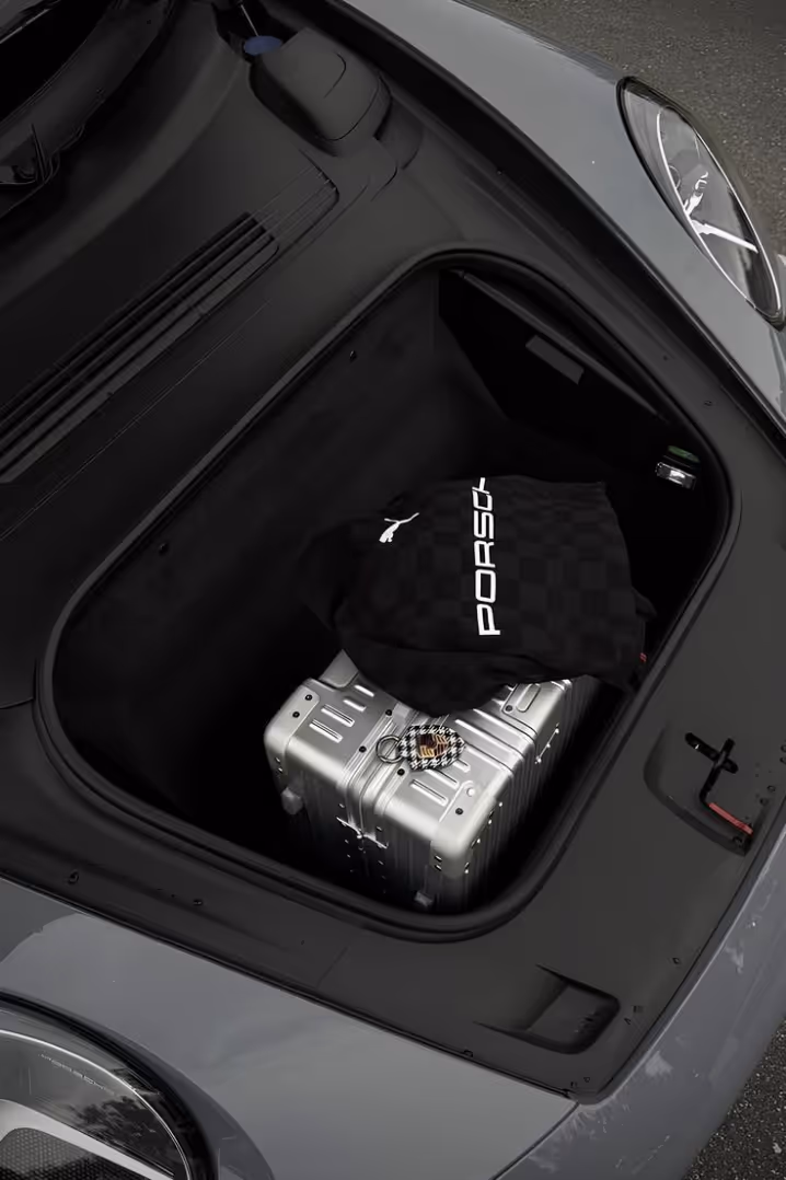 Open front trunk of a gray Porsche with silver suitcase and black Porsche Puma bag inside.