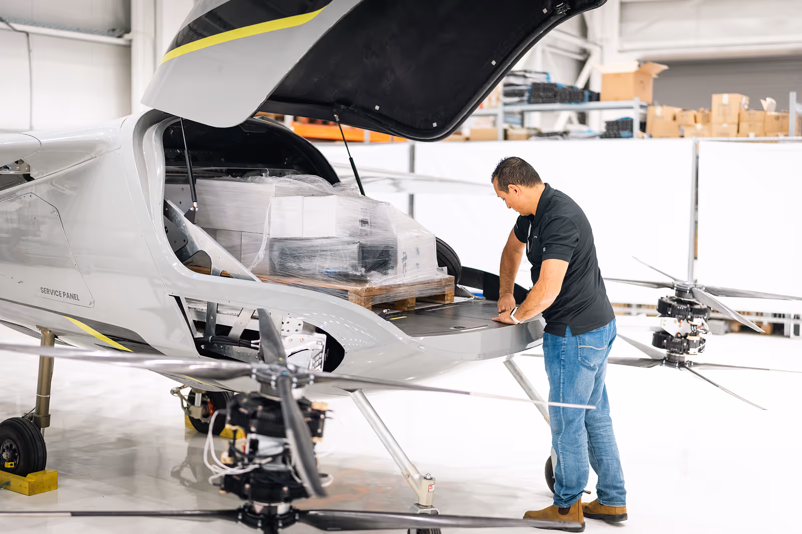 Man in black shirt inspects cargo hold of a small aircraft loaded with wrapped packages on a pallet.
