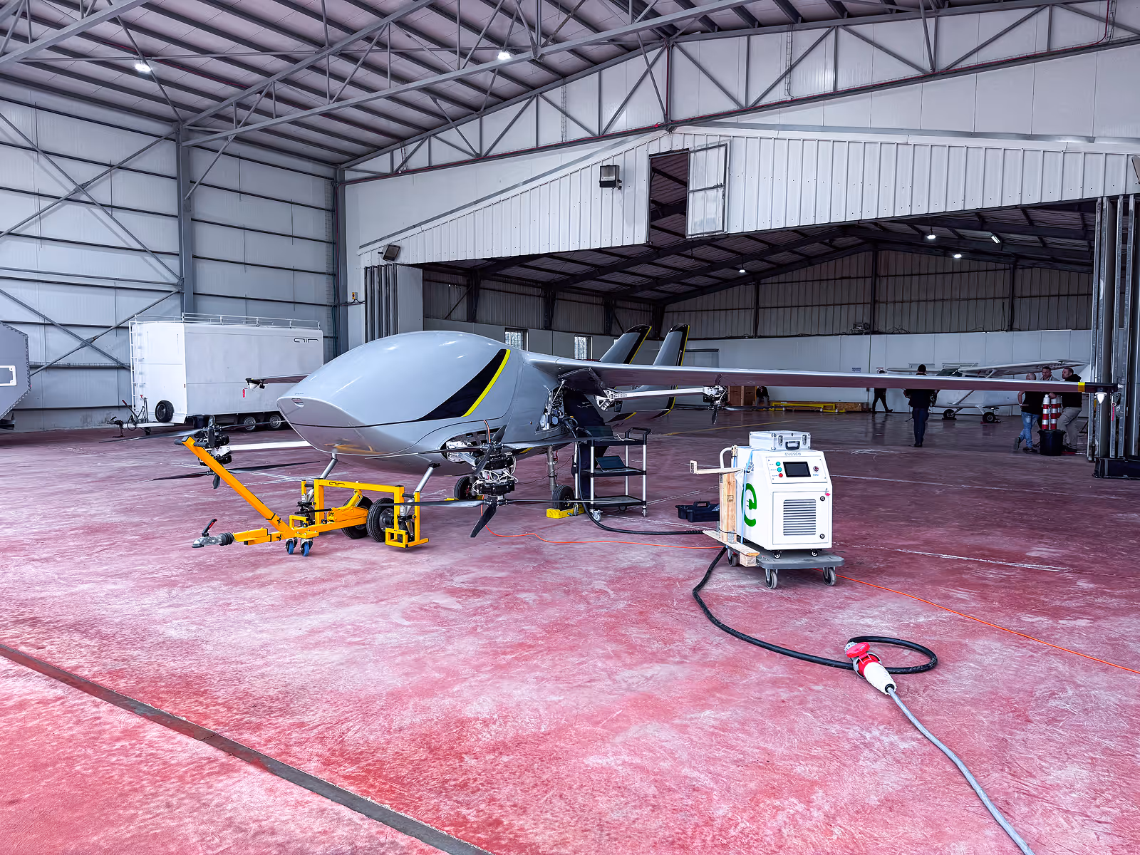 Small unmanned aircraft in a large hangar connected to a charging station with cables and equipment nearby.